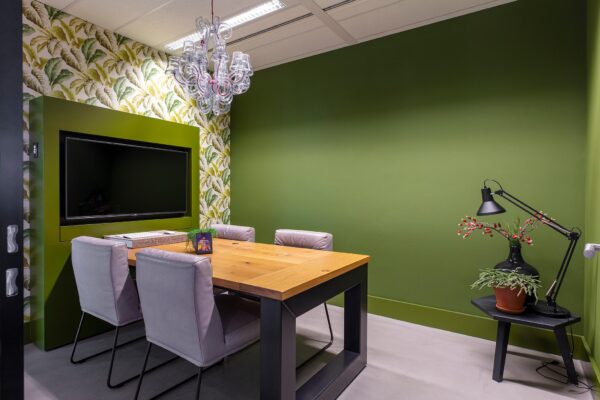 Kleine vergaderruimte met groen-geverfde muren, een houten tafel, vier stoelen en een muurtelevisie aan de Vliegend Hertlaan.
